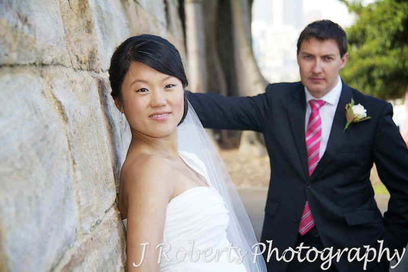 Bride looking along a wall at the camera with groom behind - wedding photography sydney